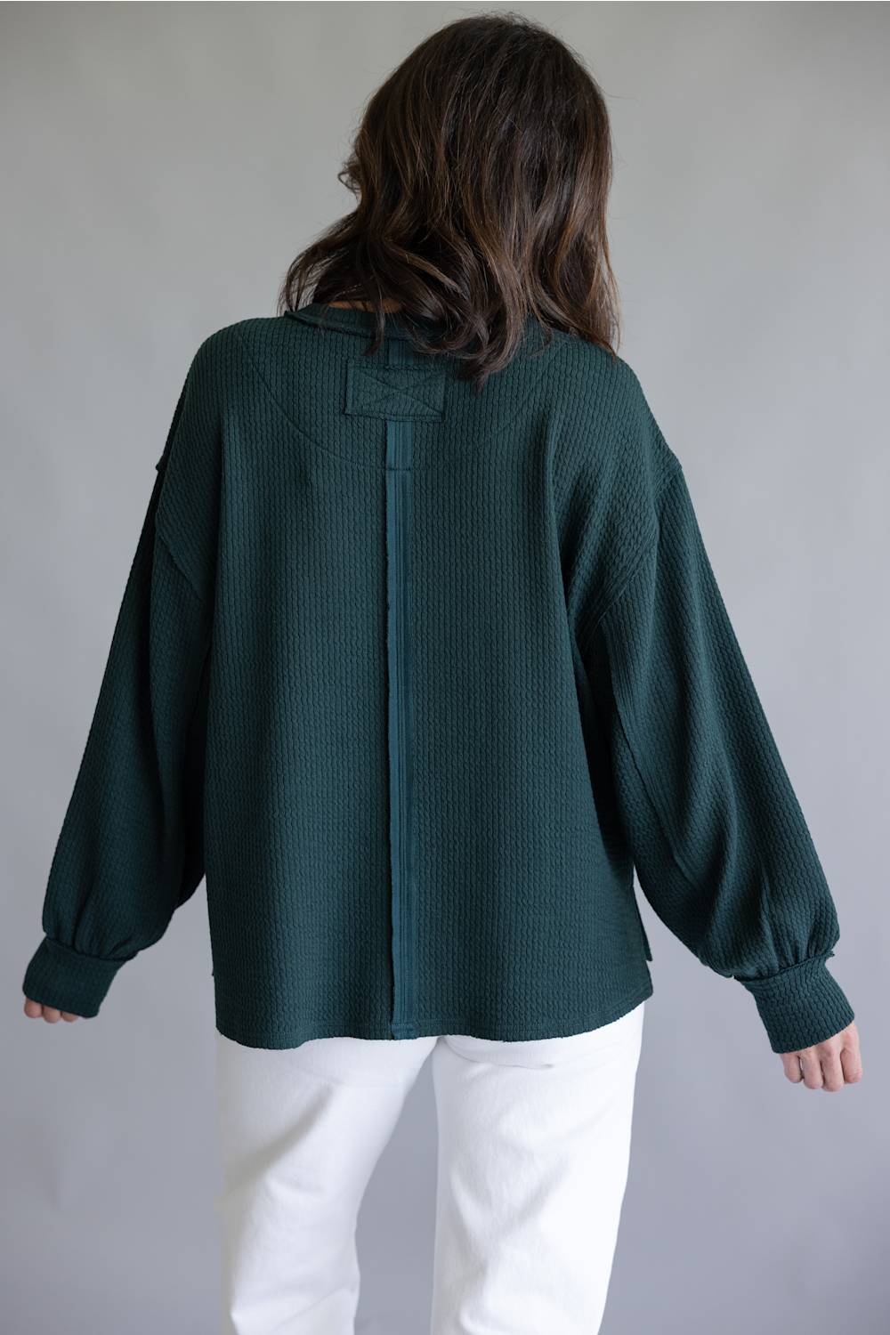 A woman viewed from the back wearing a dark forest green textured knit sweater with balloon sleeves and white pants, set against a plain gray studio background.