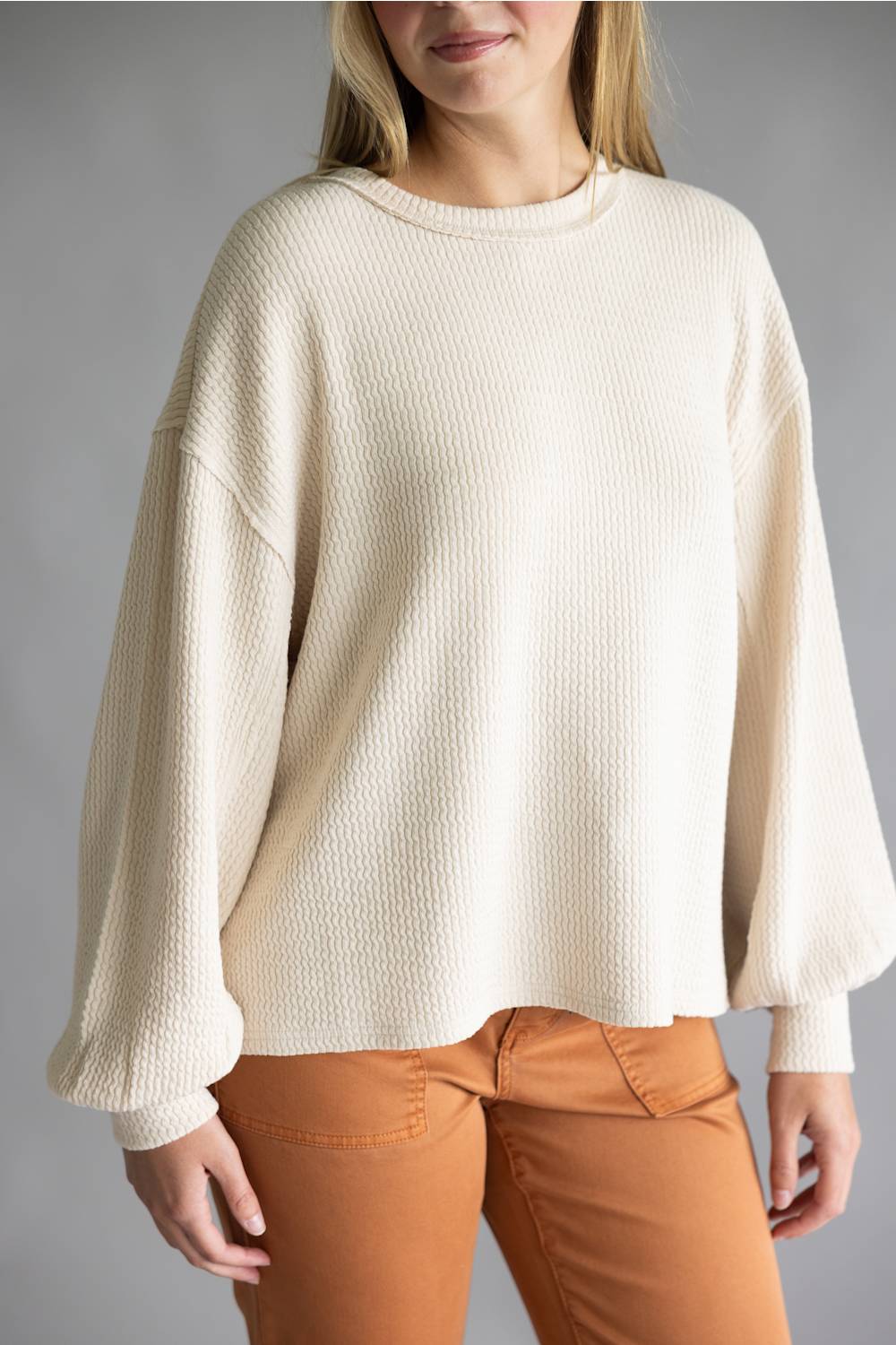 A woman wearing a cream-colored, textured waffle-knit sweater with balloon sleeves and orange-brown pants, shown from the chin down against a neutral gray background.
