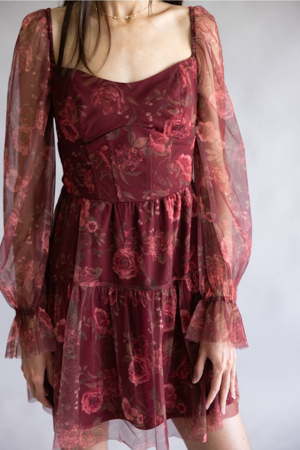 A woman wearing a burgundy floral mini dress with sheer long sleeves, a sweetheart neckline, and a tiered skirt, photographed against a plain light gray background.
