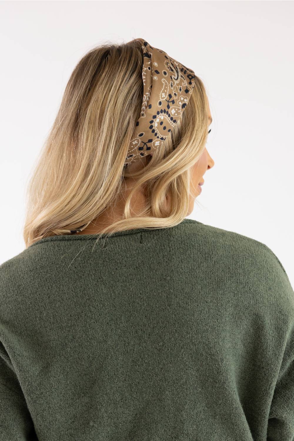 A woman shown from the back wearing a beige bandana headband with a black paisley pattern, paired with an olive green knit sweater, against a plain white background.