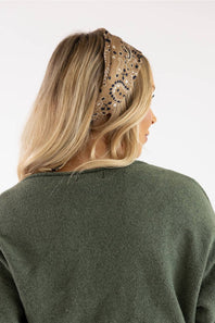 A woman shown from the back wearing a beige bandana headband with a black paisley pattern, paired with an olive green knit sweater, against a plain white background.