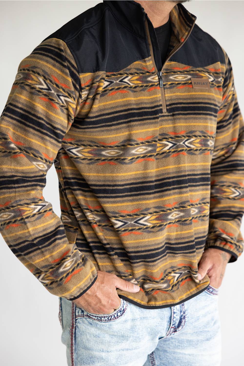 A man wearing a long-sleeve quarter-zip fleece pullover with a Southwestern geometric pattern in brown, beige, black, and orange, paired with light-wash blue jeans.