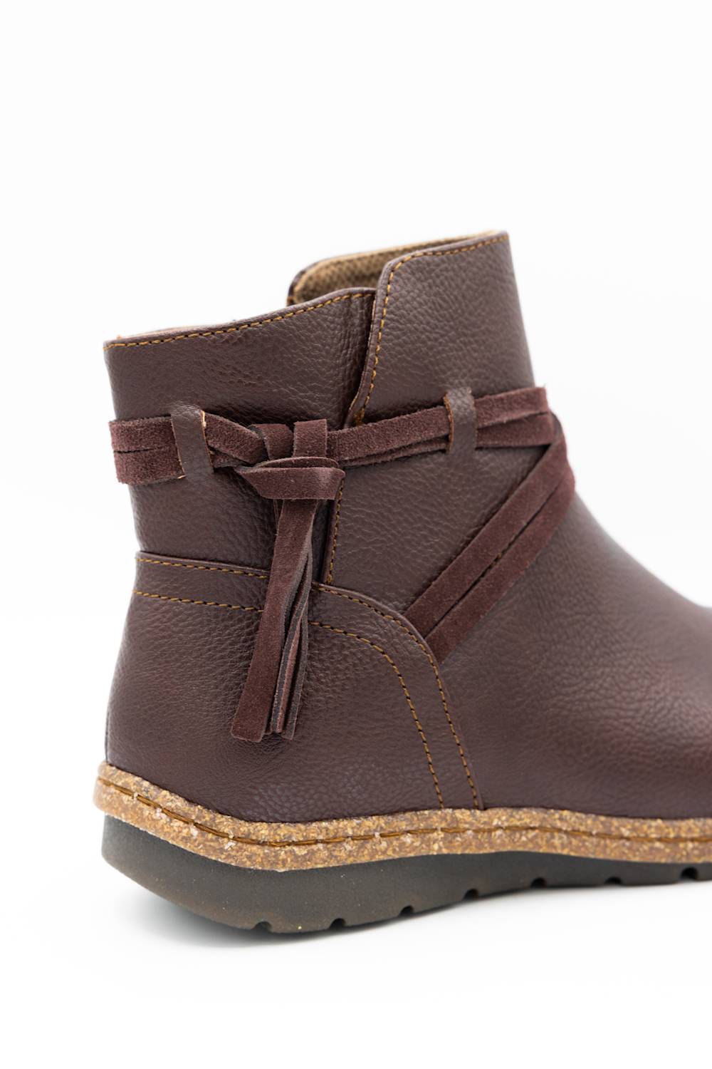 A dark brown pebbled leather ankle boot with a decorative side bow and tassel detail, a cork-wrapped midsole, and a dark gray outsole, shown against a plain white background.