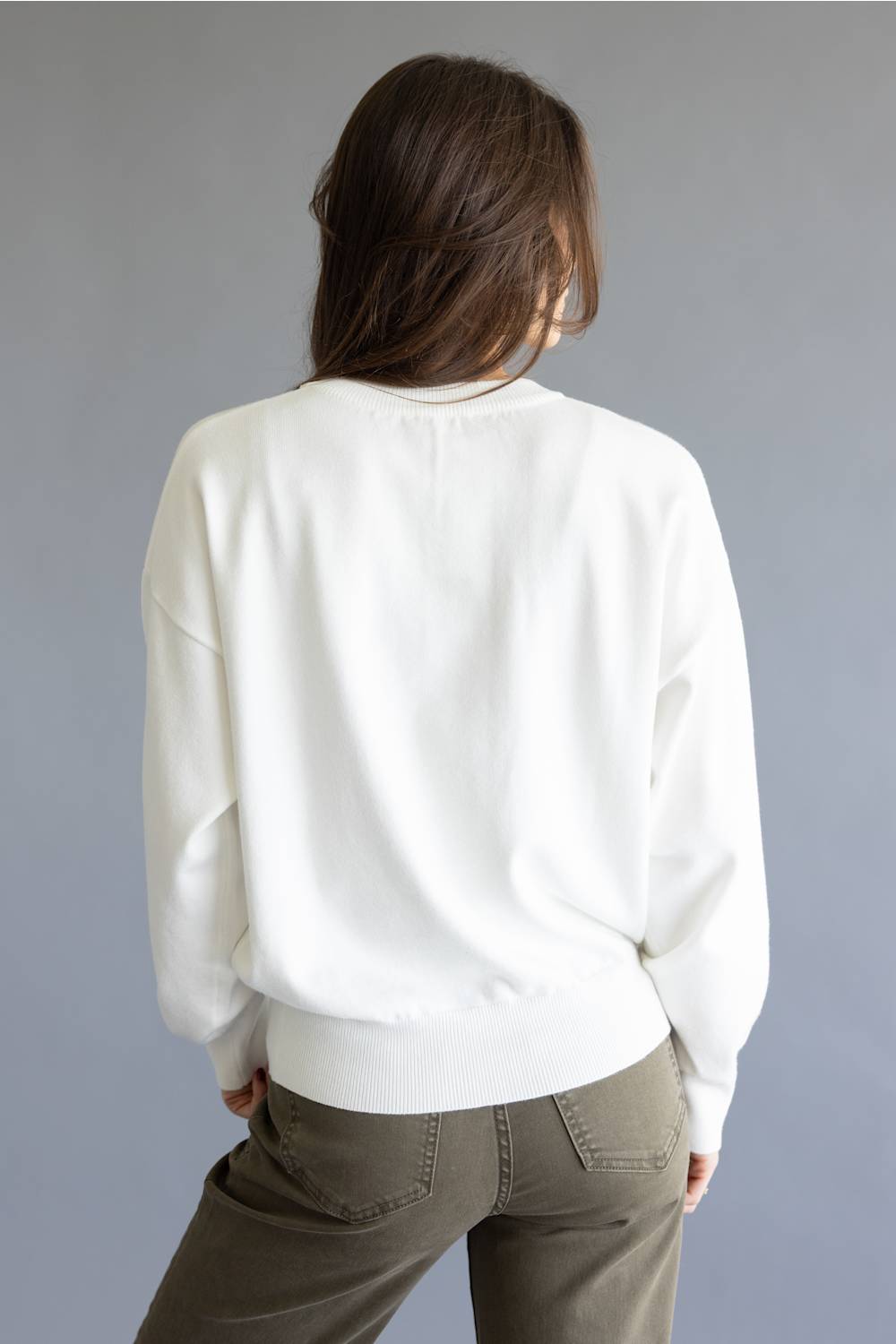 A woman viewed from behind wearing a white crewneck sweatshirt and olive green pants, set against a plain gray studio background.