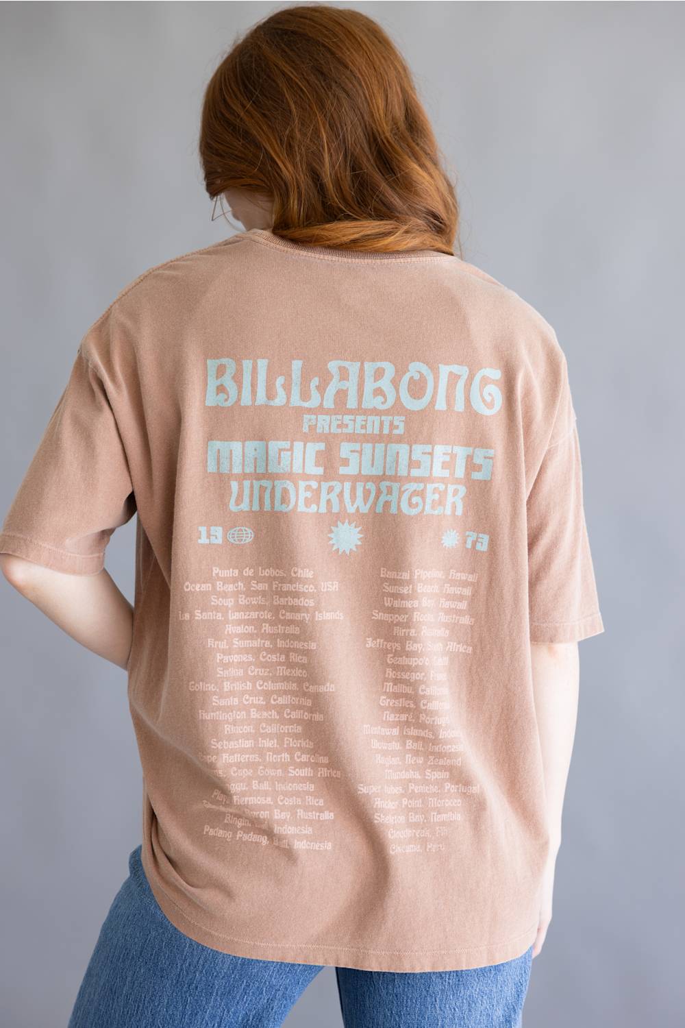 A person shown from the back wearing a light peach-pink oversized T-shirt with large pale blue and white text reading Billabong Presents Magic Sunsets Underwater, paired with blue jeans against a plain gray studio background.