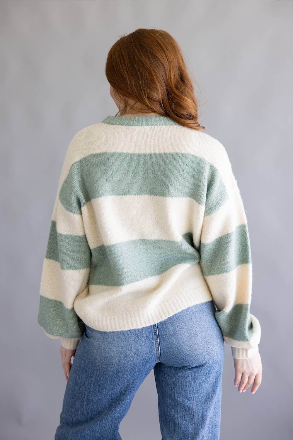 A woman viewed from behind wearing a cream and sage green striped knit sweater and blue jeans, standing against a plain gray studio background.