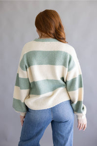 A woman viewed from behind wearing a cream and sage green striped knit sweater and blue jeans, standing against a plain gray studio background.