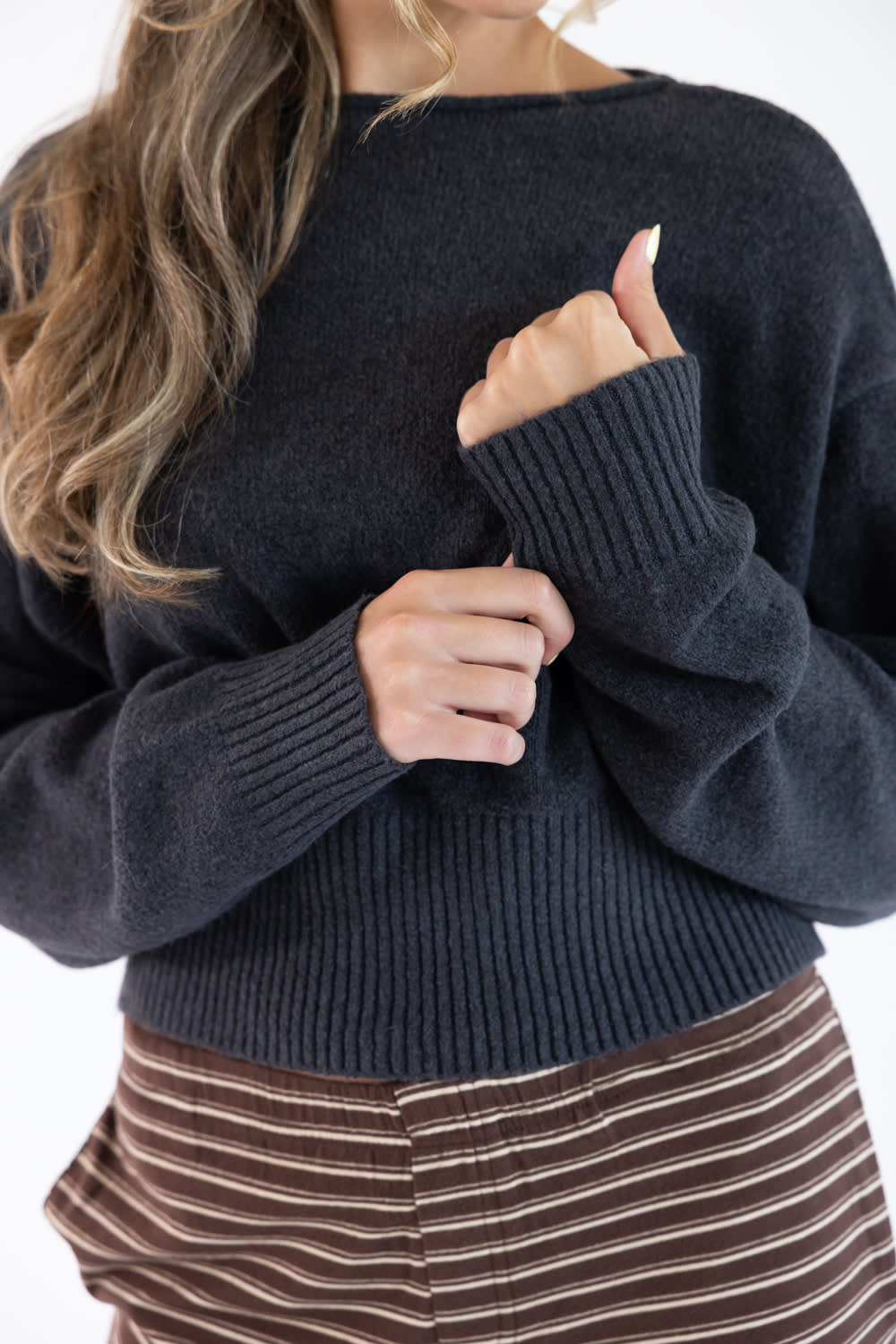 A close-up of a woman wearing a dark charcoal gray ribbed knit sweater, paired with brown and white striped pants, against a plain white background.