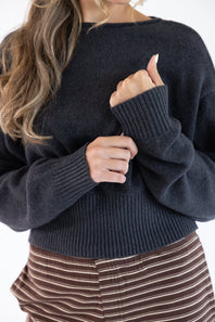 A close-up of a woman wearing a dark charcoal gray ribbed knit sweater, paired with brown and white striped pants, against a plain white background.