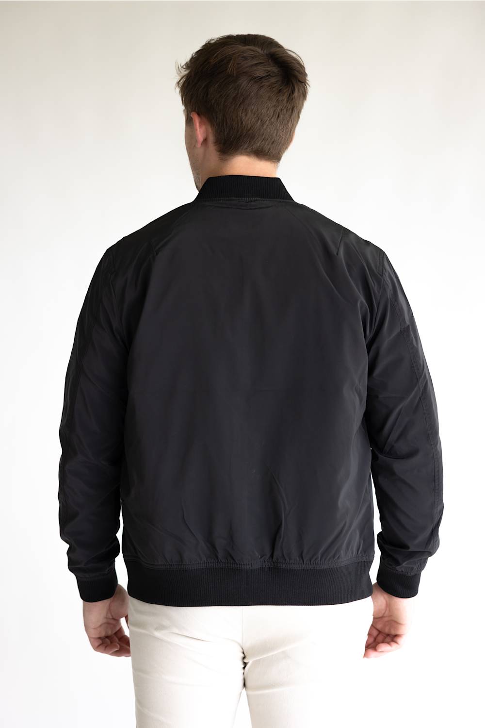 A man viewed from behind wearing a black bomber jacket and white pants, standing against a plain light background.