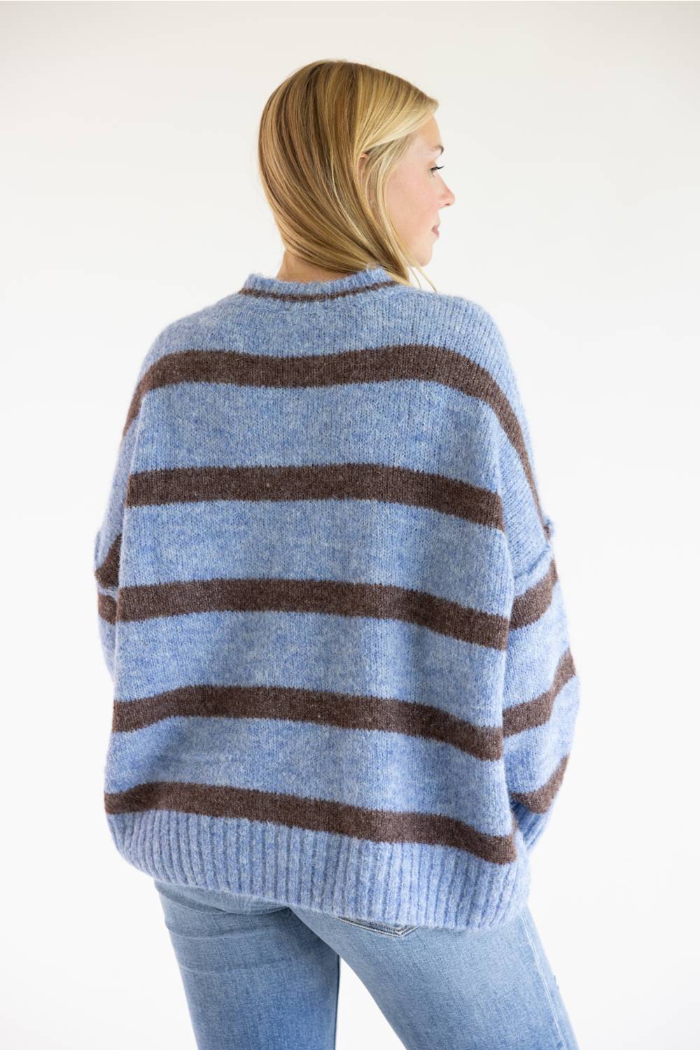 A woman shown from the back wearing a loose, oversized knit sweater with horizontal brown stripes on a light blue base, paired with light blue jeans against a plain white background.