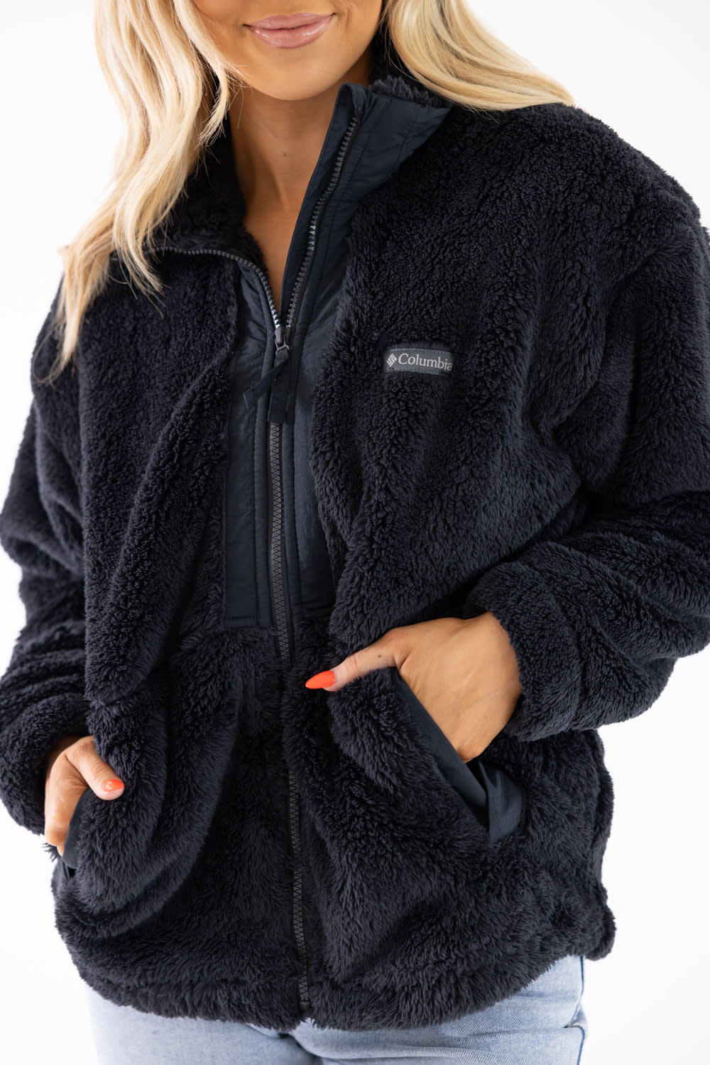 A woman wearing a black sherpa fleece zip-up jacket with a small Columbia logo patch on the chest, paired with light blue jeans against a white background.