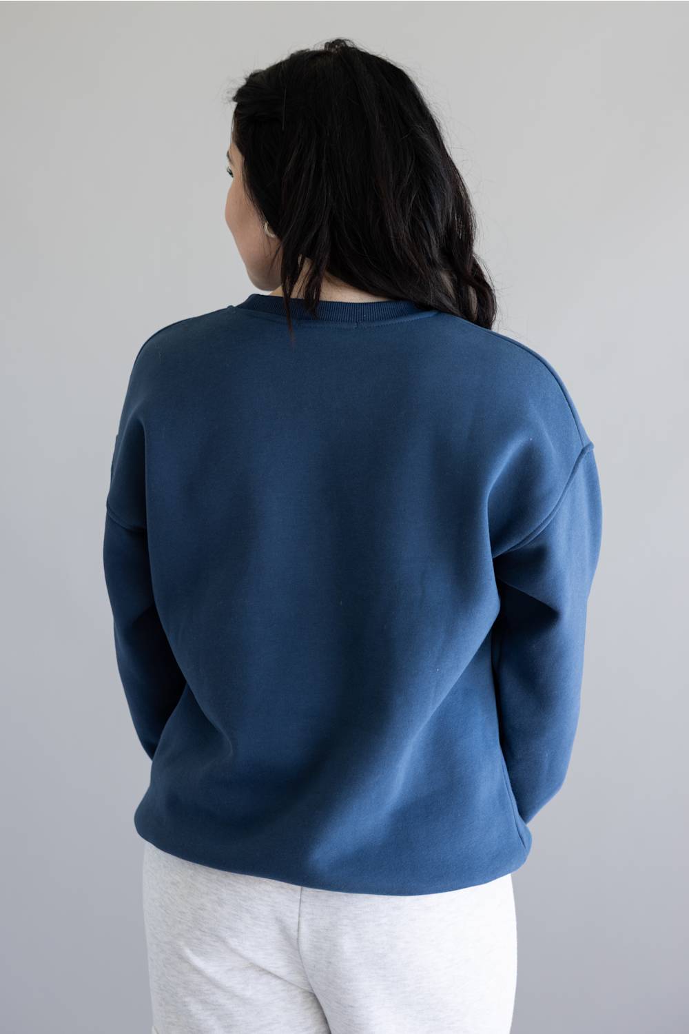 A person shown from the back wearing a dark blue crewneck sweatshirt and light gray sweatpants against a plain light gray background.