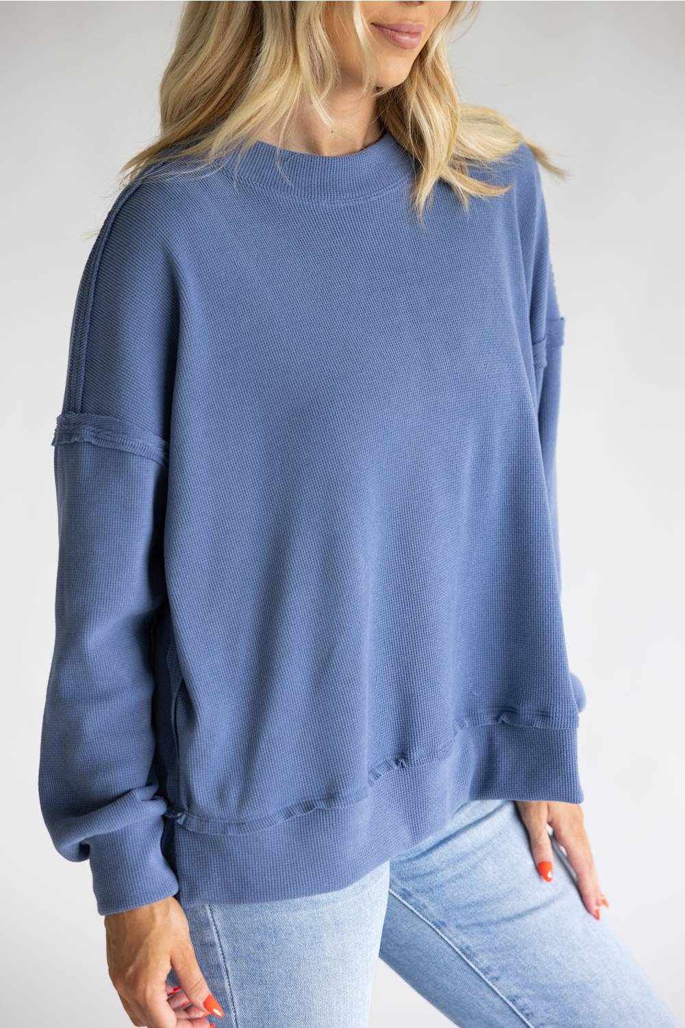 A woman wearing an oversized dusty blue crewneck sweatshirt with raw, frayed hems and sleeve details, paired with light-wash blue jeans against a plain light gray background.