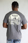 A man viewed from behind wearing a charcoal gray t-shirt with a large white circular graphic on the back featuring cacti, mountains, and the text 'BUSCH LIGHT'.