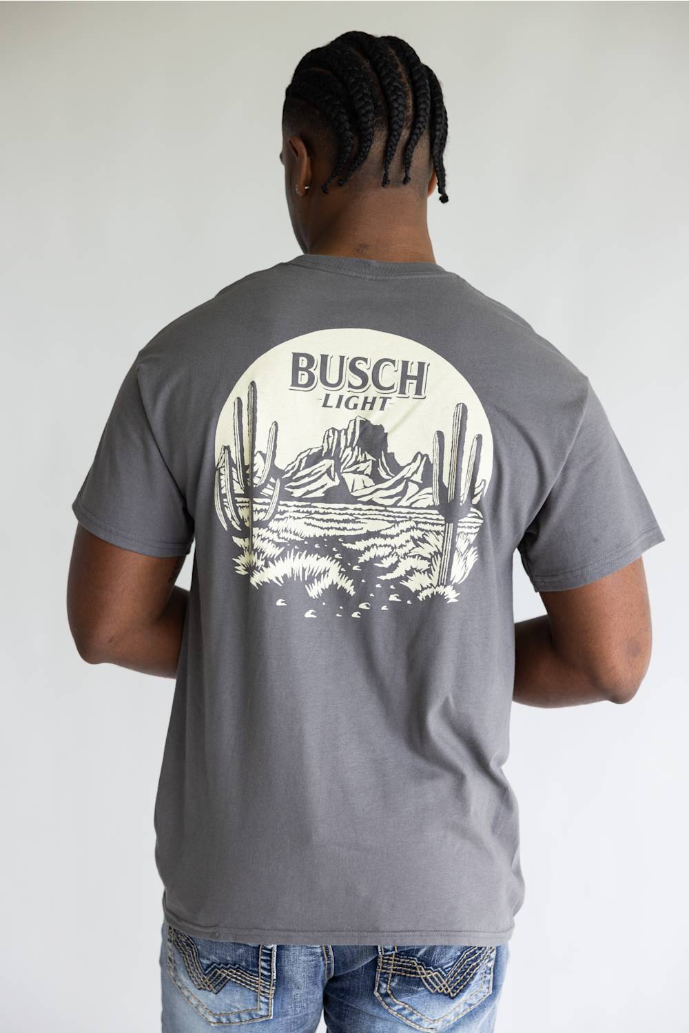 A man viewed from behind wearing a charcoal gray t-shirt with a large white circular graphic on the back featuring cacti, mountains, and the text 'BUSCH LIGHT'.