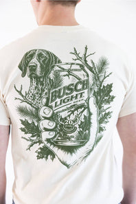 A man wearing a cream-colored t-shirt, viewed from the back. The shirt features a large green graphic print of a hunting dog, pine branches, and a hand holding a can of Busch Light beer.