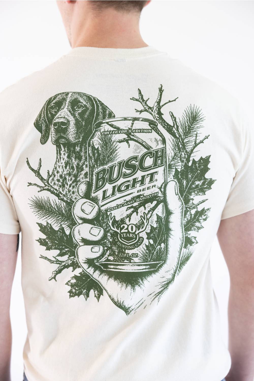 A man wearing a cream-colored t-shirt, viewed from the back. The shirt features a large green graphic print of a hunting dog, pine branches, and a hand holding a can of Busch Light beer.