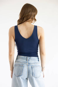 A woman viewed from behind wearing a navy blue scoop-neck tank top and light-wash blue jeans against a plain white background.