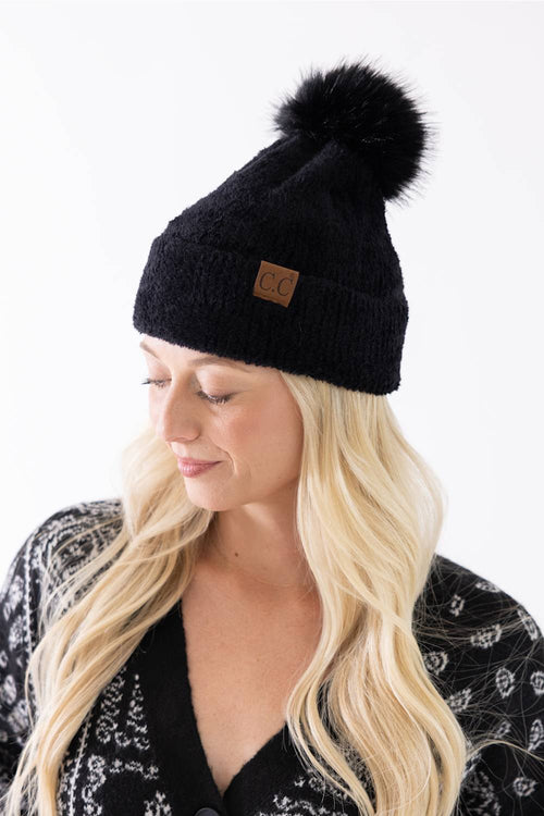 A woman wearing a black plush pom-pom beanie featuring a C.C. branded leather patch on the cuffed hem, paired with a black and white patterned cardigan.