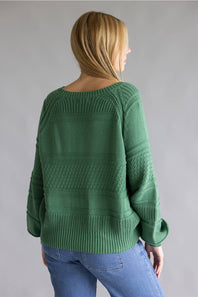 A woman viewed from the back wearing a green textured knit sweater and blue jeans against a plain gray studio background.