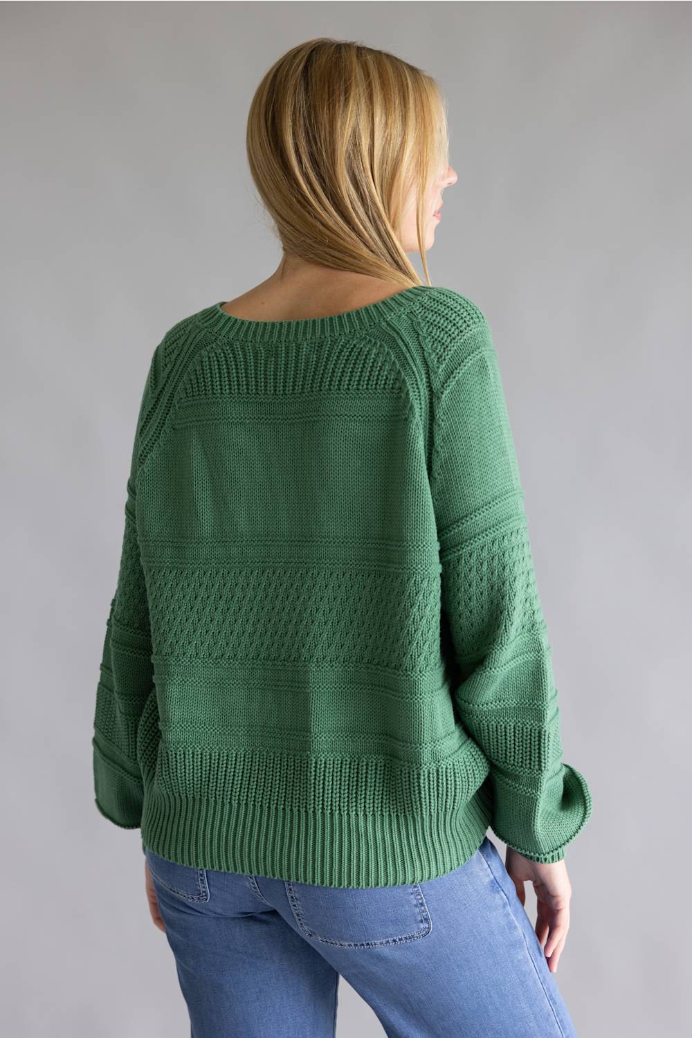 A woman viewed from the back wearing a green textured knit sweater and blue jeans against a plain gray studio background.