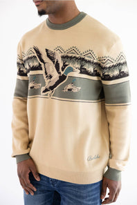 A man wearing a beige crewneck sweater with a green collar and cuffs, featuring a graphic of a mallard duck and mountains across the chest, paired with blue jeans.