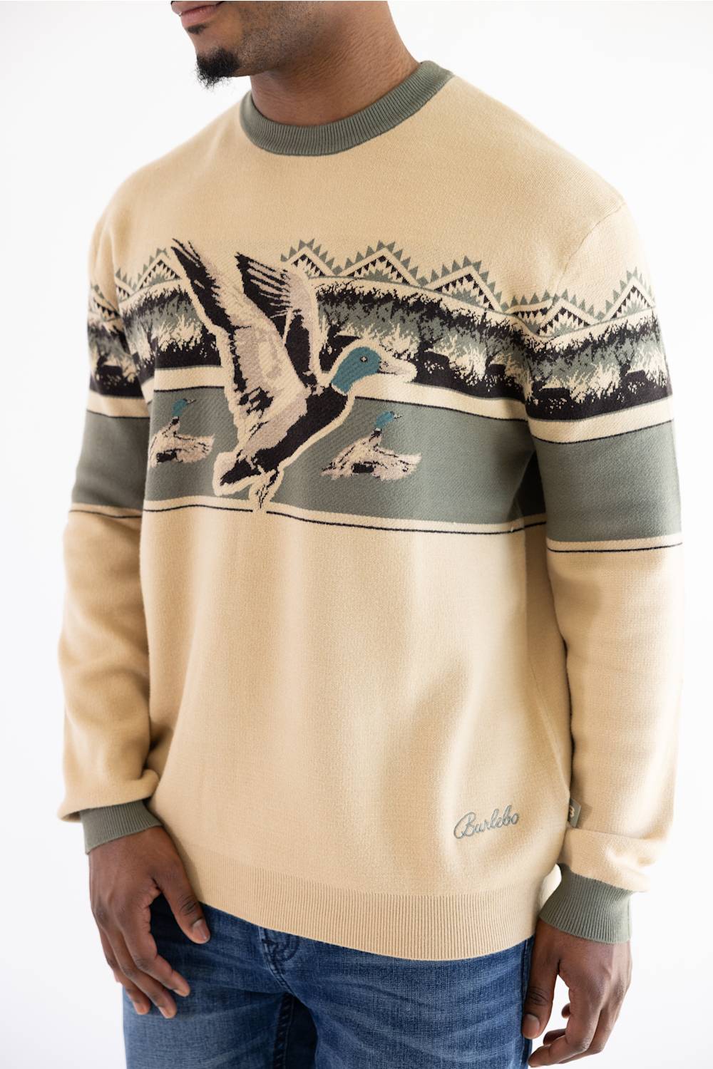 A man wearing a beige crewneck sweater with a green collar and cuffs, featuring a graphic of a mallard duck and mountains across the chest, paired with blue jeans.