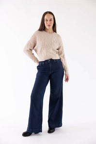A woman modeling a beige cable-knit sweater and dark blue wide-leg jeans against a plain white studio background.