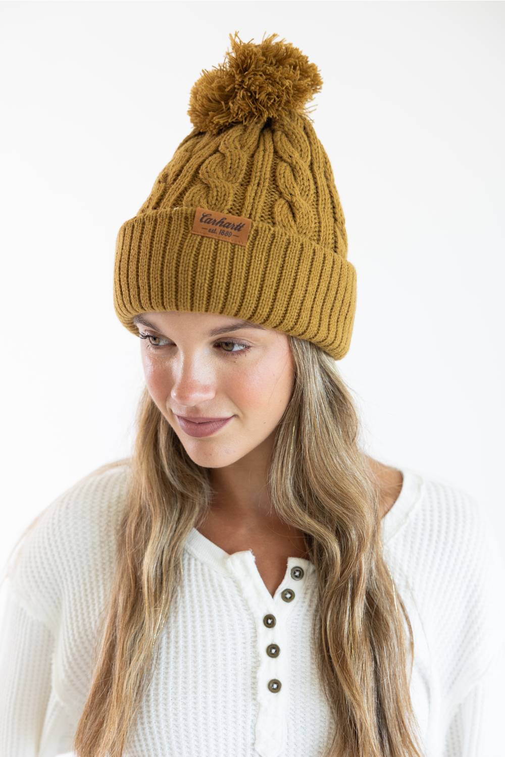 A woman wearing a mustard yellow cable-knit beanie with a brown pom-pom and a small brown brand patch on the front, paired with a white henley top against a plain white background.