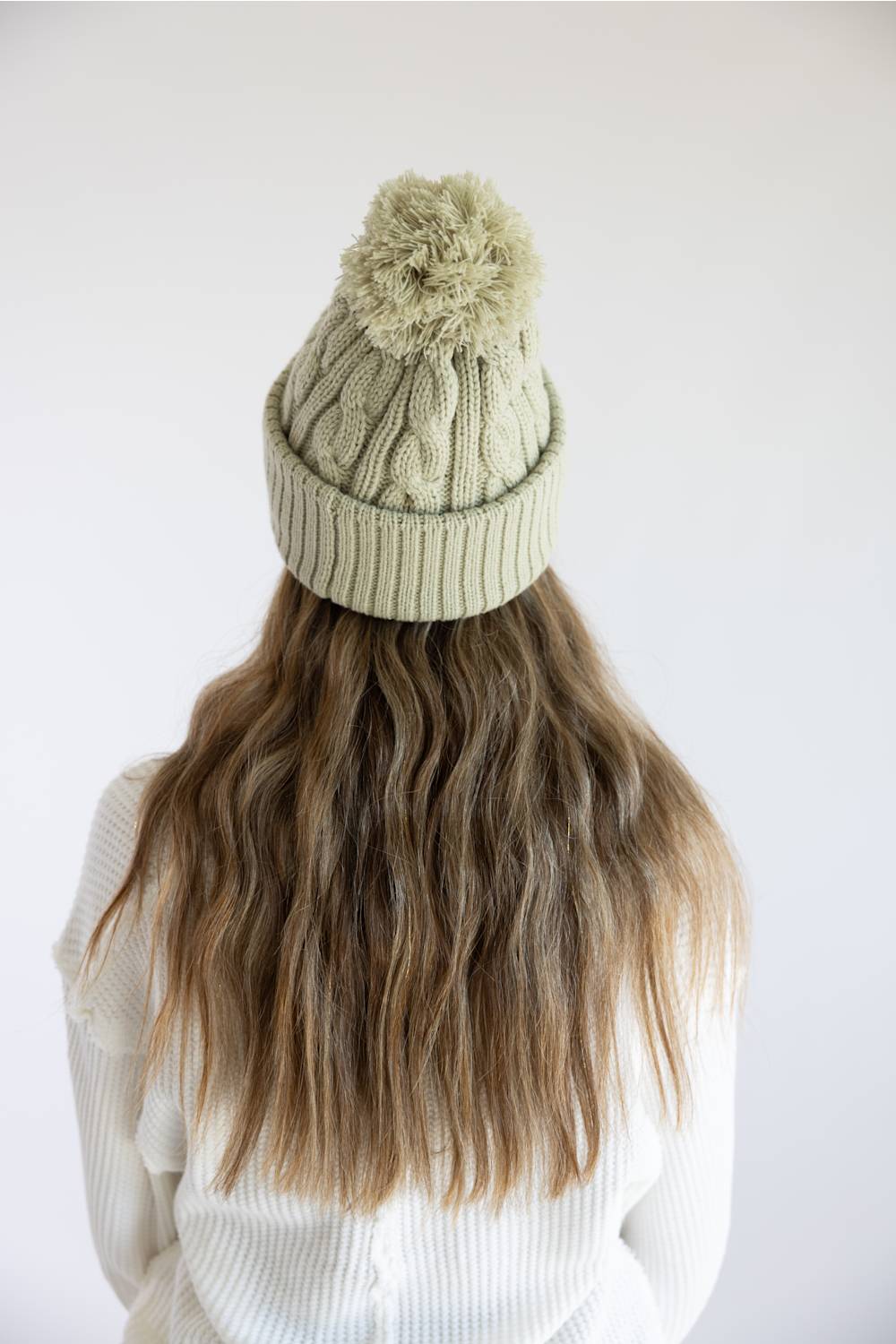 A person viewed from behind wearing a light sage green cable-knit beanie with a matching pom-pom, paired with a white ribbed sweater against a plain white background.