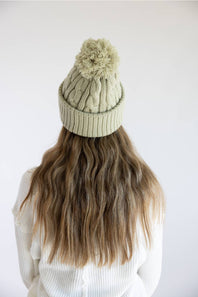 A person viewed from behind wearing a light sage green cable-knit beanie with a matching pom-pom, paired with a white ribbed sweater against a plain white background.