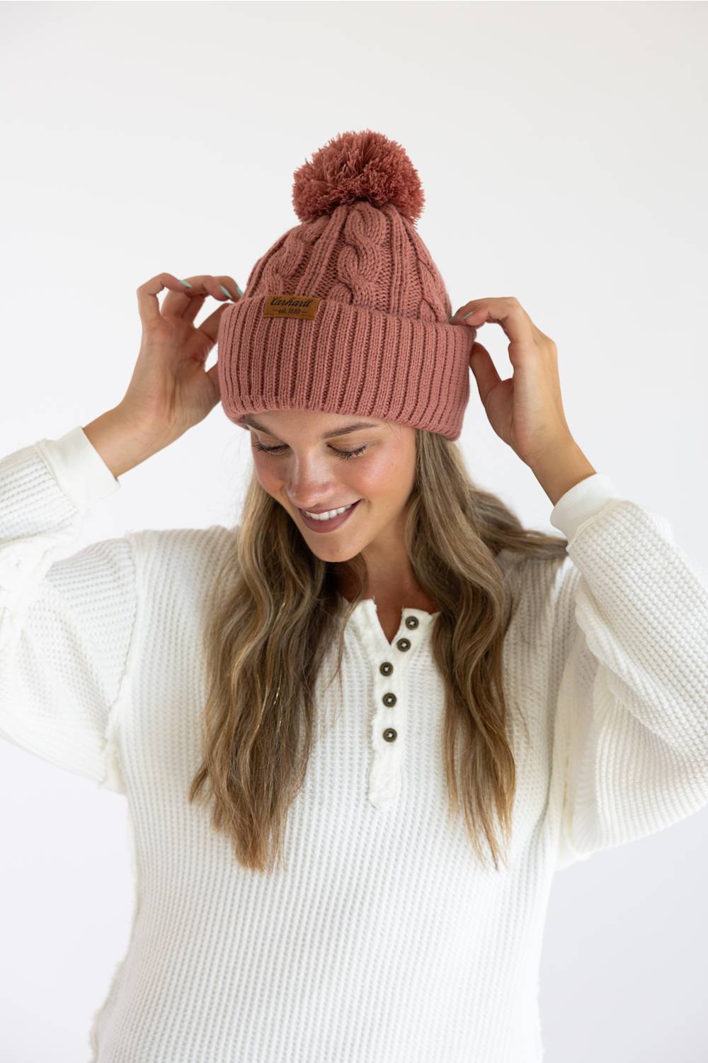 A woman wearing a dusty rose pink cable-knit beanie with a matching pom-pom and a small brown leather brand patch, posing in a white ribbed henley sweater against a clean white background.