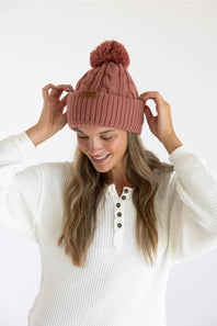 A woman wearing a dusty rose pink cable-knit beanie with a matching pom-pom and a small brown leather brand patch, posing in a white ribbed henley sweater against a clean white background.