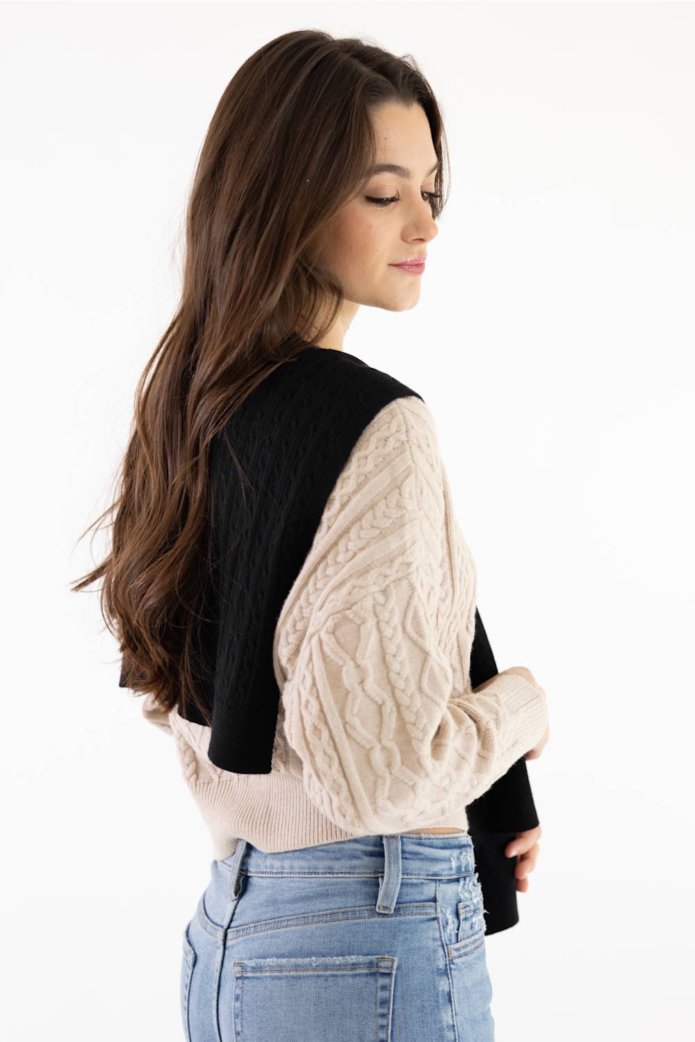 A woman shown from the back wearing a color-block knit sweater with a black yoke and sleeves, and a cream cable-knit lower body, paired with light blue jeans against a white studio background.