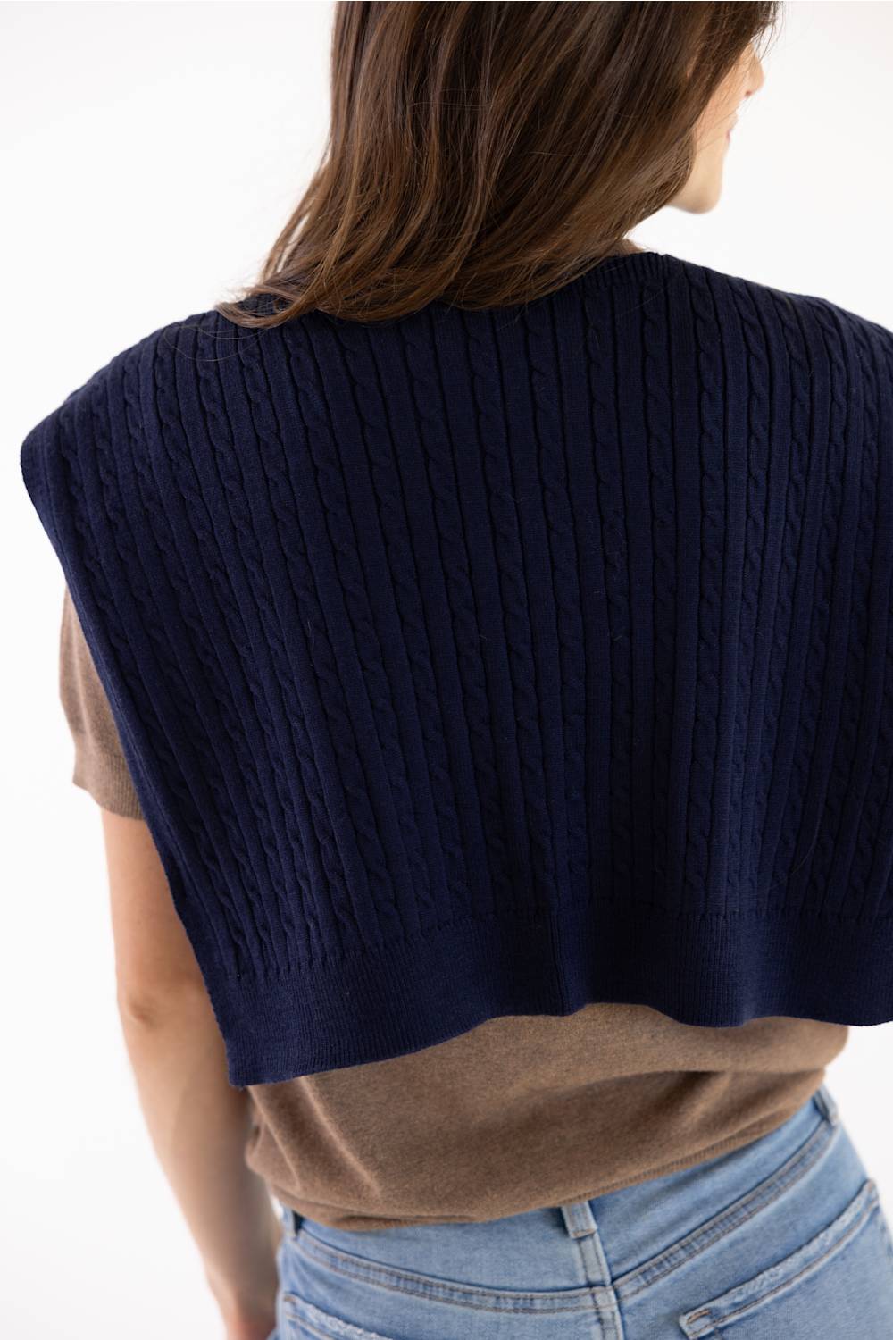 A woman shown from the back wearing a navy blue cable-knit cropped shrug or cape over a brown t-shirt and light blue jeans, against a plain white background.