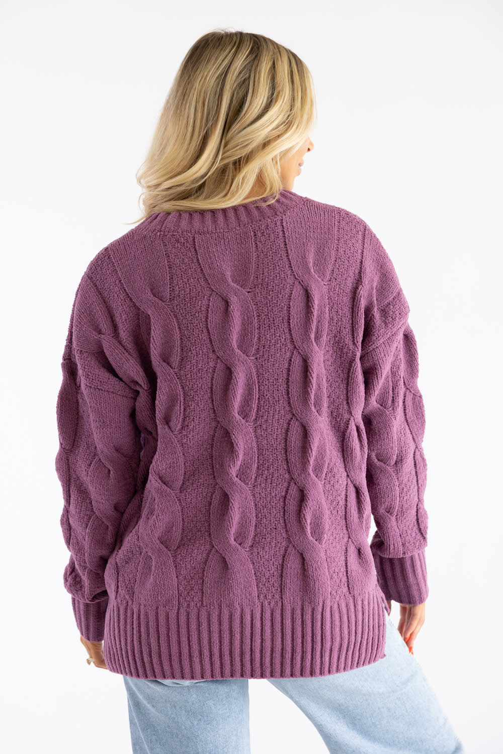 A woman viewed from behind wearing an oversized cable-knit sweater in a dusty mauve-purple color, paired with light blue jeans against a plain white background.