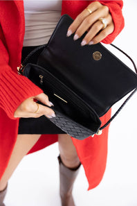 A close-up of a person holding an open black clutch handbag with a woven black quilted exterior and gold-tone hardware, wearing a bright red cardigan and black skirt against a white background.