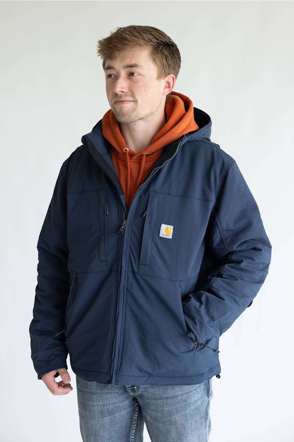A man wearing a navy blue Carhartt jacket over an orange hoodie and blue jeans, standing against a plain light gray background.