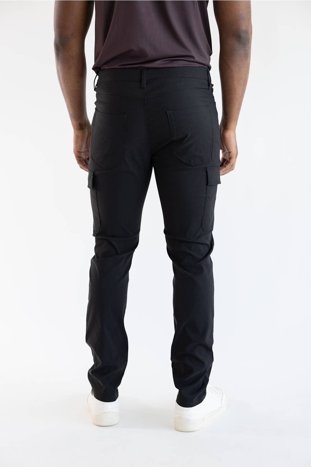 Rear view of a person wearing black cargo pants, a dark brown t-shirt, and white sneakers against a plain white studio background.