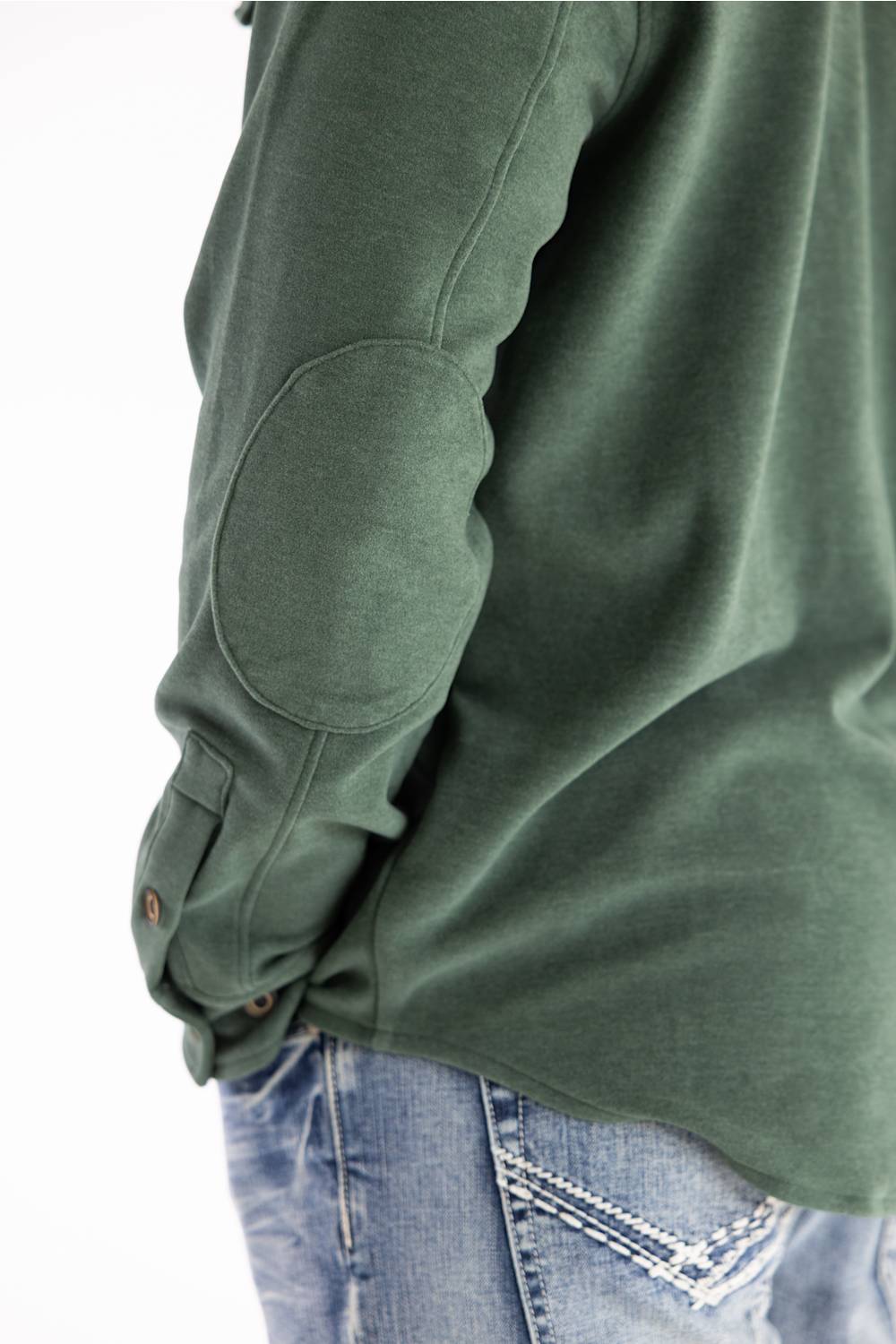 Close-up of a person wearing an olive green long-sleeve top with elbow patches and buttoned cuffs, paired with blue jeans against a white background.