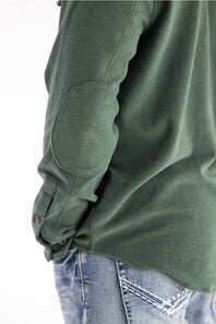 Close-up of a person wearing an olive green long-sleeve top with elbow patches and buttoned cuffs, paired with blue jeans against a white background.