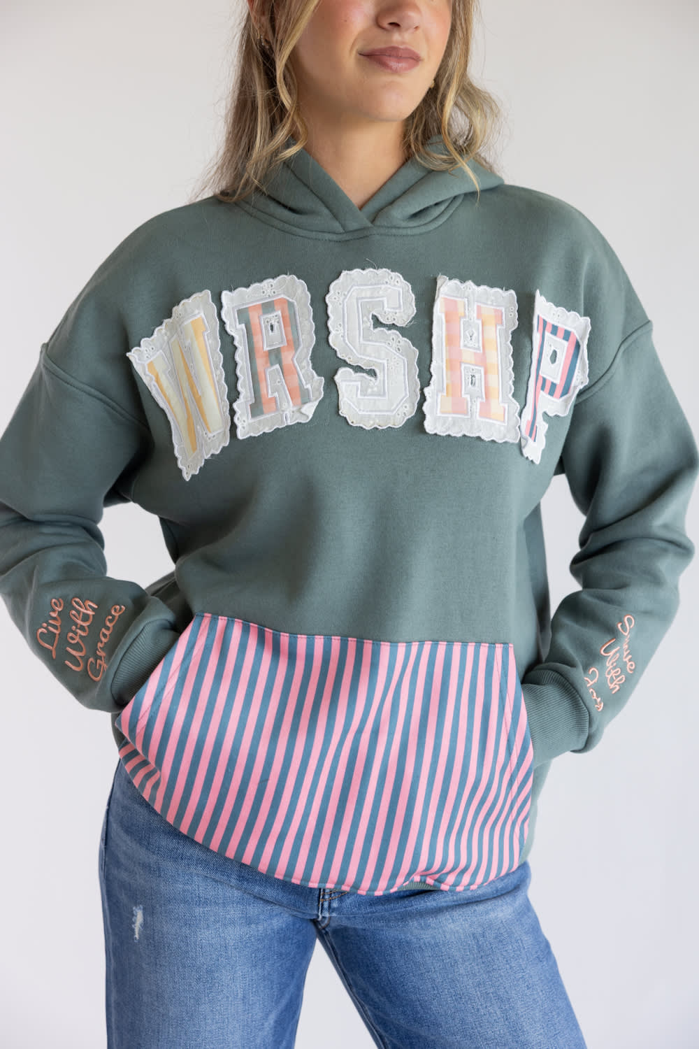 A woman wearing a sage green hoodie with a large 'WRSHP' graphic across the chest in white patches with pastel lettering, paired with a pink and blue striped shirt tucked into the hoodie, and blue jeans.
