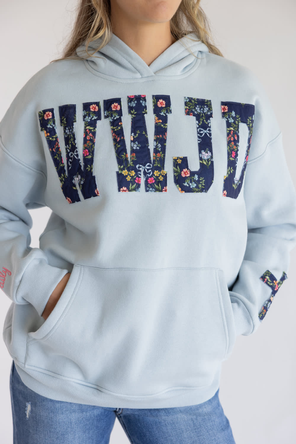 A person wearing a light blue pullover hoodie with large floral-patterned letters spelling 'WILD' across the chest, paired with blue jeans against a plain light background.