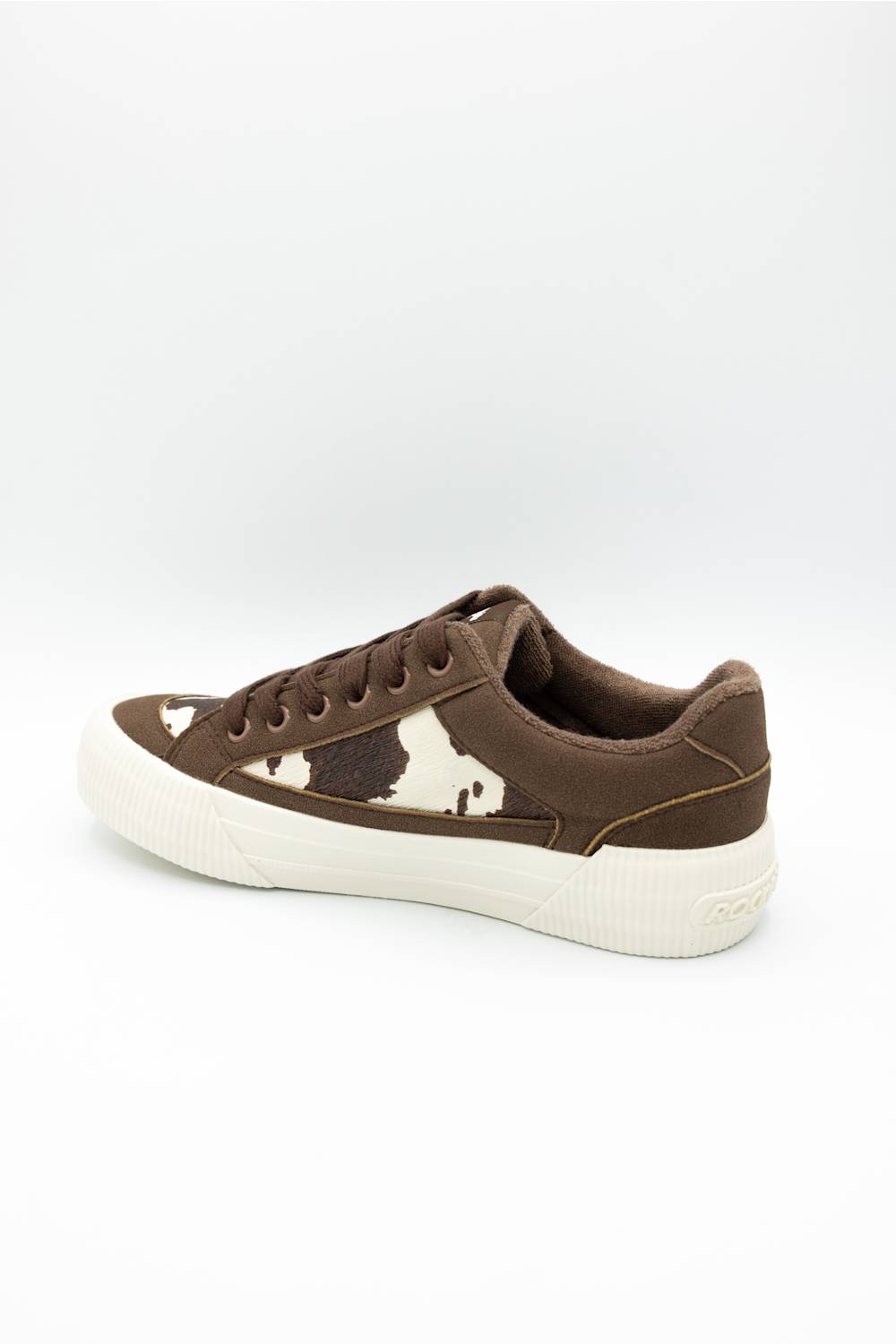 A low-top sneaker in brown suede with a brown and white cow-print pattern on the side and a thick white rubber sole, shown from a side profile on a white background.
