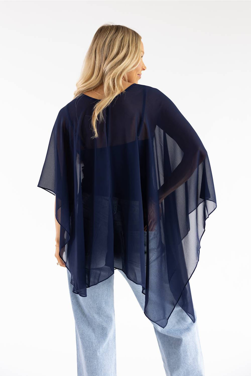 A woman shown from the back wearing a sheer navy blue poncho-style top over a light blue top and jeans, against a plain white studio background.