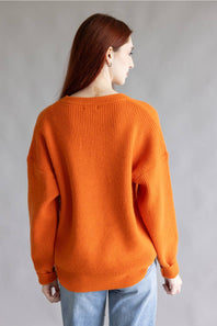 A woman with long auburn hair is shown from the back wearing an oversized orange ribbed knit sweater and light blue jeans against a plain gray studio background.