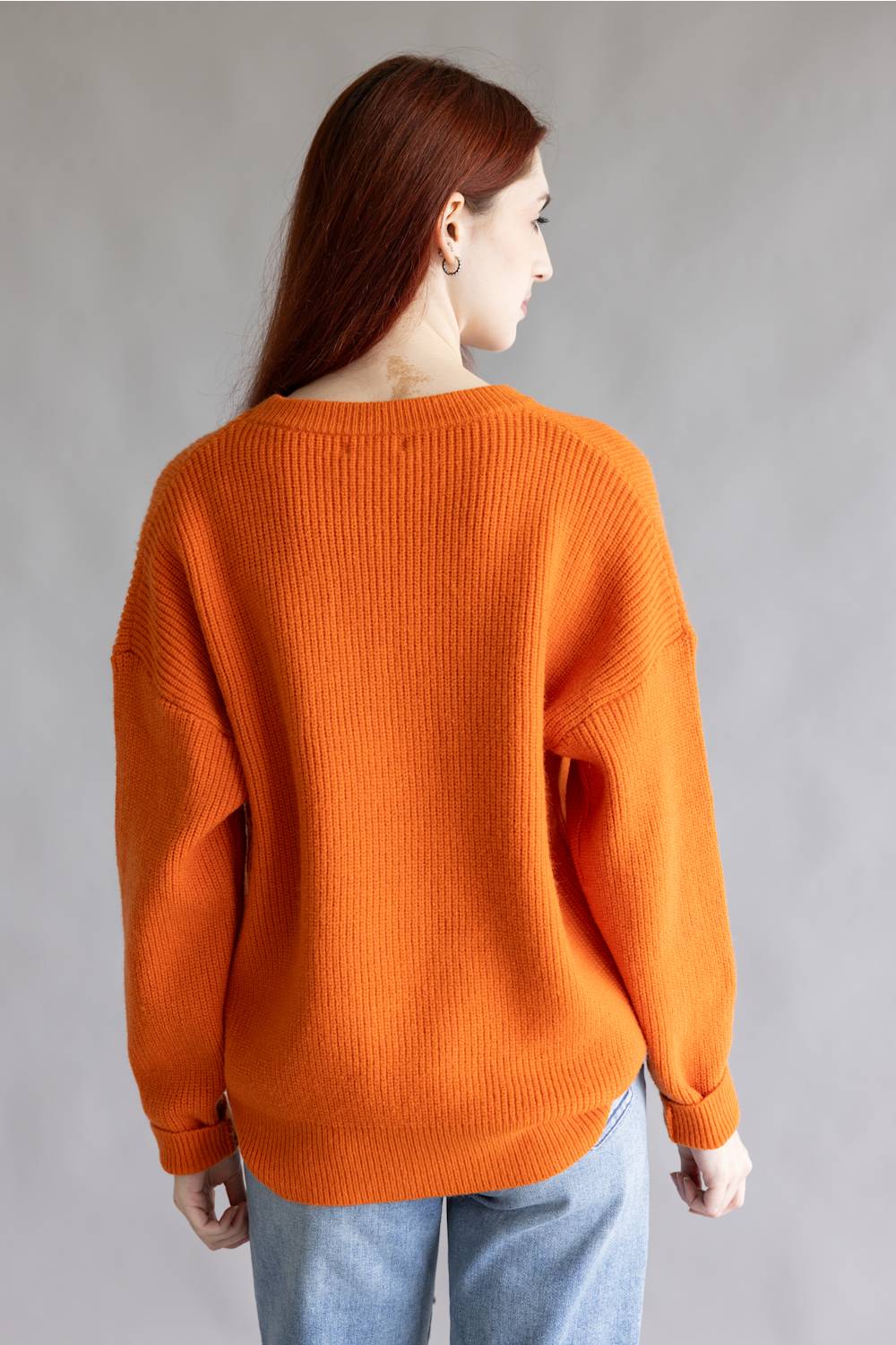 A woman with long auburn hair is shown from the back wearing an oversized orange ribbed knit sweater and light blue jeans against a plain gray studio background.
