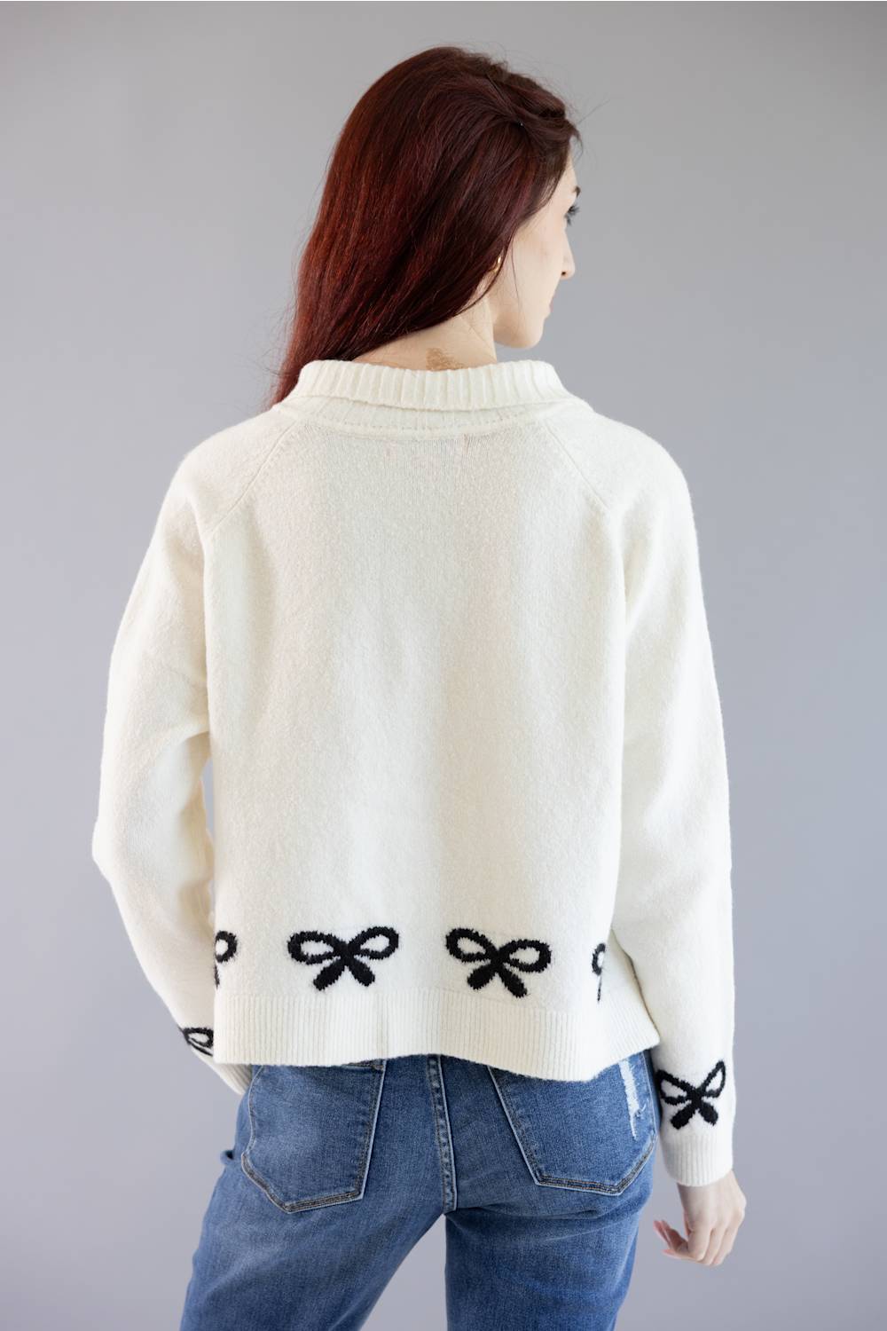 A woman viewed from the back wearing a cream-colored knit sweater with black bow details along the hem and cuffs, paired with blue jeans, against a solid gray studio background.