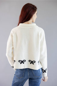 A woman viewed from the back wearing a cream-colored knit sweater with black bow details along the hem and cuffs, paired with blue jeans, against a solid gray studio background.